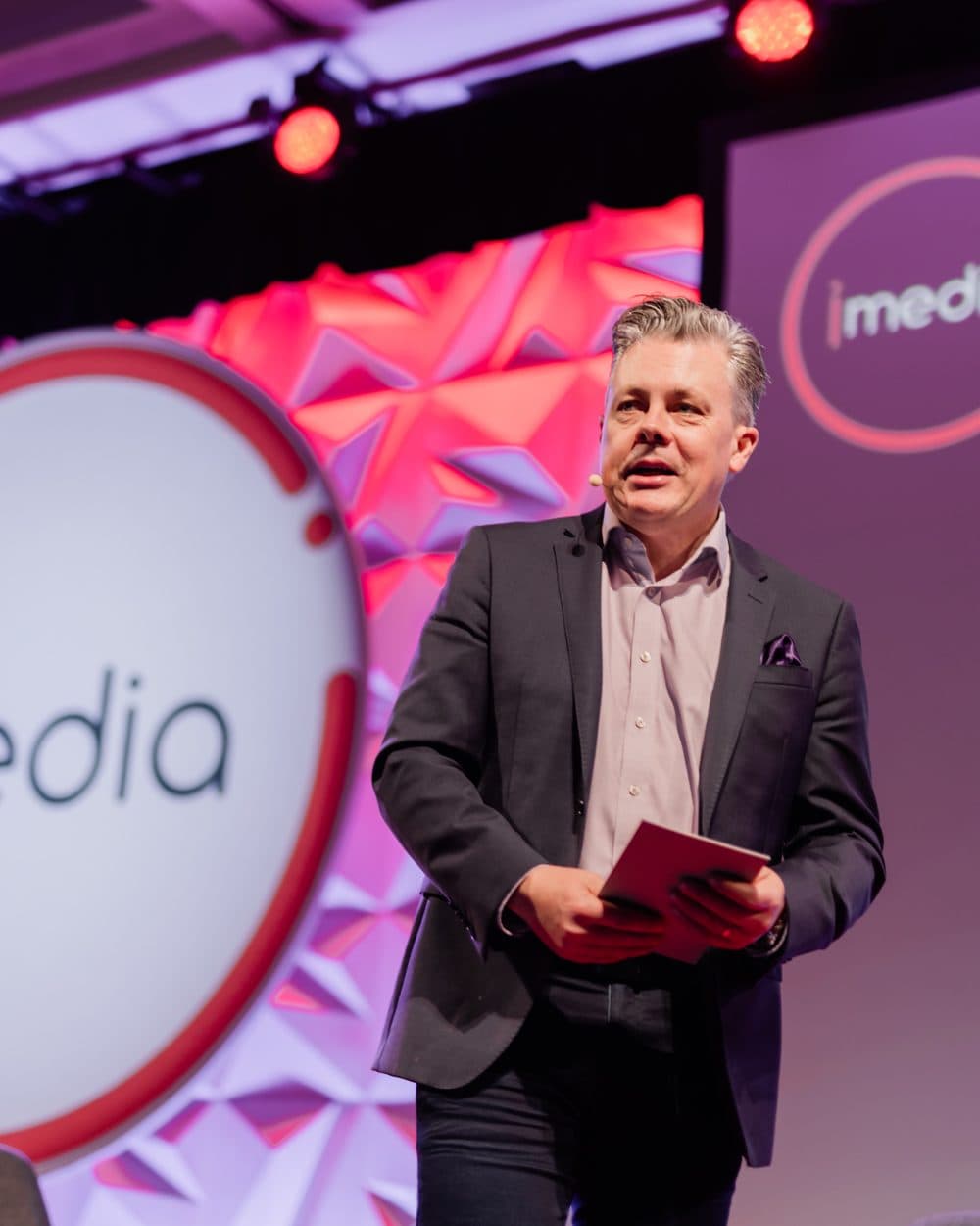 Mark Jones hosting from the stage, host cards in hand, walking toward the room mid-sentence